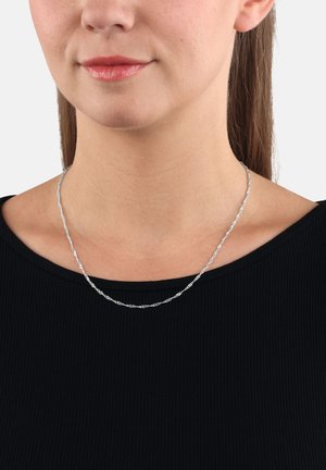 Donna che indossa una collana d'argento intrecciata sopra un top nero a coste, mostrando il collo, il mento e le labbra con un rossetto rosa chiaro.