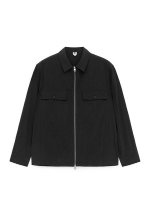 Veste noire à manches longues avec col pointu, fermeture éclair à l'avant, et deux poches poitrine à rabat avec boutons, présentée à plat sur un fond blanc.