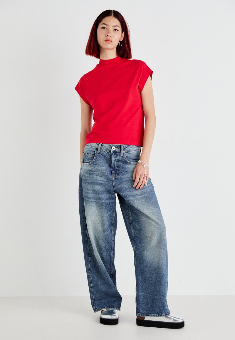 Czerwona bawełniana bluzka z krótkim rękawem i stójką, w komplecie z jasnoniebieskimi spodniami o szerokich nogawkach z wyblakłym wykończeniem i pięcioma kieszeniami. Czarne buty na platformie.