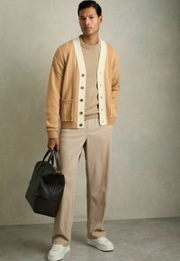 Cardigan en laine beige avec des accents et des boutons crème, associé à un pantalon en maille beige et des baskets blanches, tenant un sac en cuir noir.