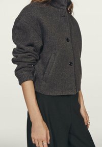 Veste en laine mélangée gris foncé avec un col haut, fermeture à boutons, poches latérales et manches légèrement oversize. Texture lisse, design décontracté.