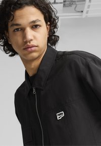 Juodas zip-up džemperis, lengvas audinys, smailas apykaklė, mažas kairės krūtinės kišenė su balta Puma logo. Lygus paviršius, laisvas siluetas, minimalistiškas dizainas.