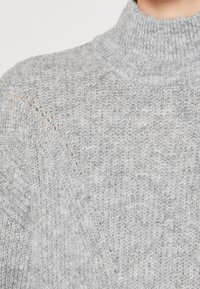 Grå stickad tröja med hög ribbad krage, med ett texturerat mönster och subtila variationer i trådfärgen genom hela plagget.