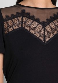 Černý top s krátkým rukávem, který má průhlednou krajkovou část se zvlněným designem kolem výstřihu a hladkým bavlněným materiálem.