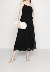 Schwarzes Spaghettiträgerkleid, plissierter Rock, leichtes Material, kombiniert mit klaren Absatzsandalen und einer weißen Clutch.