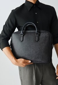 Maletín de cuero negro con asas redondeadas, cierre de cremallera y logotipo grabado. Textura lisa y forma rectangular, diseñado para uso profesional.