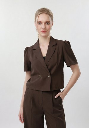 Femme blonde aux cheveux attachés porte un blazer à manches courtes marron foncé croisé et un pantalon assorti taille haute, debout devant un fond blanc.