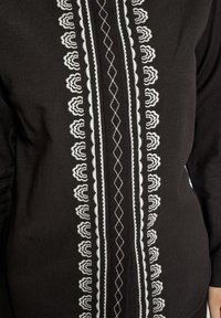 Čierny odev s prepracovanou bielou výšivkou zobrazujúcou kvetinové a diamantové vzory pozdĺž prednej stredu, zvýrazňujúci textúru a kontrast.