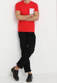 Czerwona bawełniana koszulka z białymi detalami kieszeni, zestawiona z czarnymi, podwiniętymi spodniami i czarnymi butami na sznurówki. Minimalistyczny design.