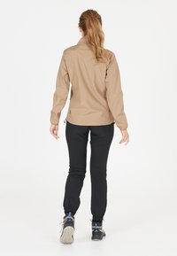 Lichtgewicht tan jacket met een hoge kraag, elastische manchetten en een trekkoord aan de onderkant, gecombineerd met fitted zwarte broek en wandelschoenen.