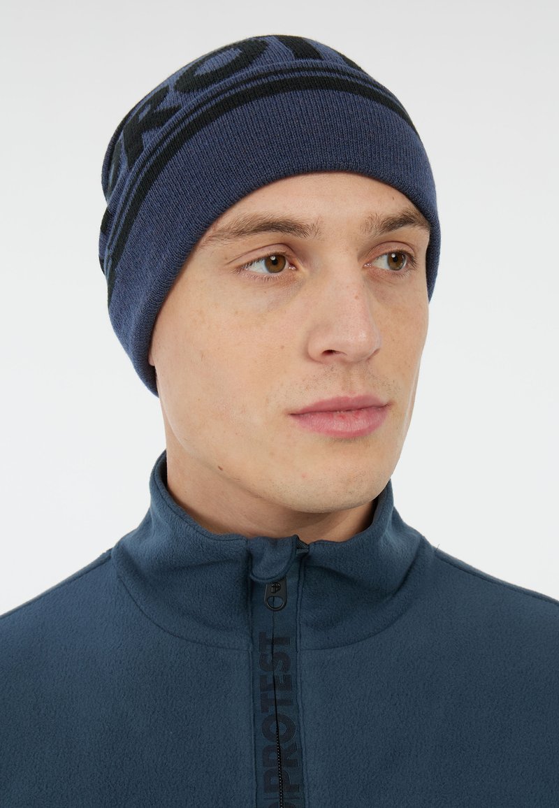 Navy brei-beanie met zwarte logoband. Het materiaal lijkt zacht en heeft een close-fitting ontwerp. Draagt met een ritsfleecejack.