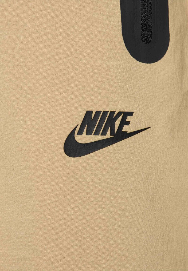 Nike beiger Stoff mit glatter Textur, versehen mit einem auffälligen schwarzen Logo-Design. Enthält einen kurzen schwarzen Reißverschluss an der Oberseite.