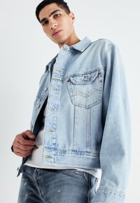 Chaqueta de mezclilla azul clara con cuello, bolsillos en el pecho y cierres de botón. Presenta áreas desgastadas y acentos de costura en contraste.