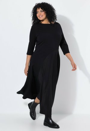 Femme aux cheveux bouclés portant une robe noire fluide à manches trois-quarts et des bottines noires, marchant sur un fond clair uni.
