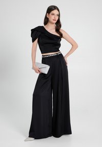 Top in satin nero a spalla unica abbinato a pantaloni a gamba larga, accentuato da una cintura con gioielli. Tiene una pochette argento, caratterizzata da una superficie piatta e liscia.