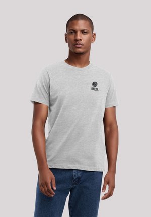 Graues Baumwoll-T-Shirt mit rundem Halsausschnitt, kurzen Ärmeln und einem schwarzen Smiley-Gesicht-Graphic auf der linken Brust. Getragen mit blauen Denim-Jeans.
