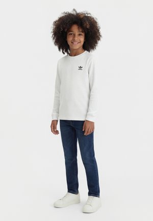Uśmiechnięte dziecko z kręconymi włosami, ubrane w białą, długorękawową koszulkę Adidas, niebieskie dżinsy i białe trampki, stojące na jednolitym tle.