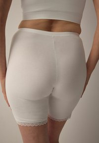 Fehér pamut rövidnadrág sima textúrával és csipke díszítéssel az alján. Testhezálló, középderékpánttal tervezve.
