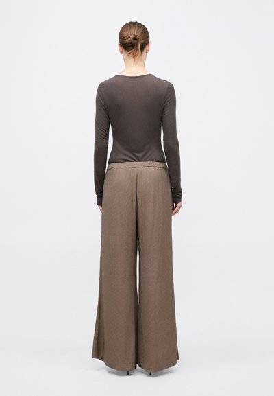 Donna con capelli raccolti in uno chignon, indossa una maglia marrone aderente a maniche lunghe e pantaloni larghi e morbidi di colore marrone chiaro, in piedi di spalle.