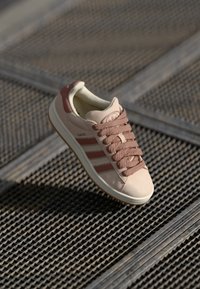 Light pink sneaker with brown accents and three side stripes. Features a textured upper, thick laces, and a rubber sole.