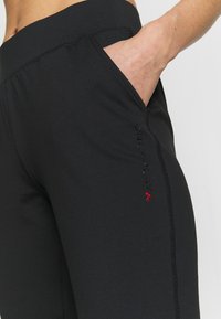 Czarne legginsy sportowe z gładką fakturą, elastycznym materiałem, boczną kieszenią oraz czerwonym logo na lewym boku.