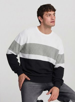 Giovane uomo seduto, indossa un maglione a righe bianco, grigio e nero, guarda verso destra con un'espressione neutra su uno sfondo semplice.