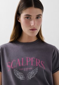 T-shirt en coton gris avec le texte rose "SCALPERS" et un graphique d'aile blanche. Manches courtes et design à col rond.