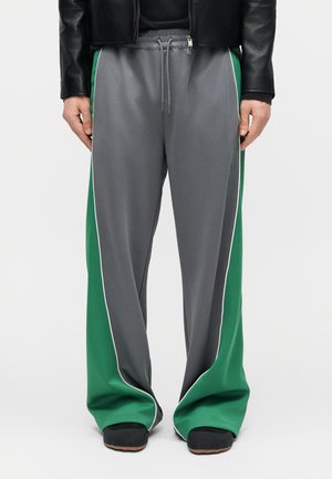 Persona che indossa pantaloni della tuta grigi larghi con pannelli laterali verdi e bordature bianche, abbinati a scarpe nere e una giacca nera.