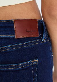 Sötétkék farmernadrág, amelyen egy barna bőr folt található, rajta a "Paga Jeans London" felirattal, amely bele van nyomva. Kontrasztos varrás és zseb részletek láthatók.