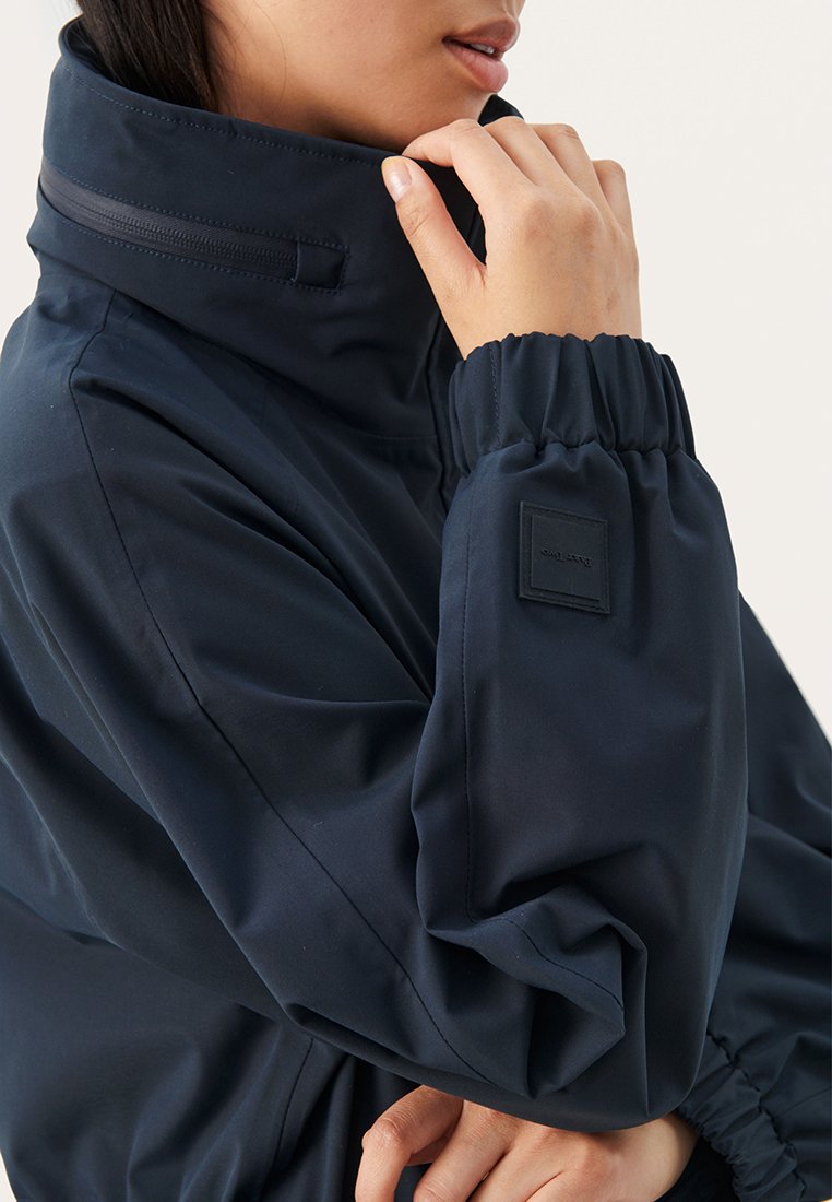 Giacca impermeabile blu navy con colletto alto, polsini elastici e una toppa quadrata con logo sul braccio. Texture liscia e vestibilità oversize.