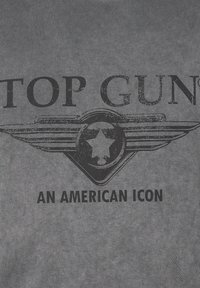 Harmaa t-paita, jossa on mustat tekstit "TOP GUN" ja "AN AMERICAN ICON" sekä keskellä siipien ja tähden logo, kaikki kuluneen tyylin mukaisia.