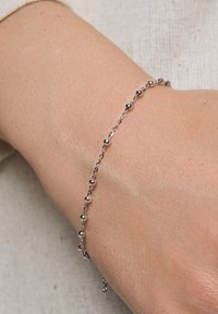 Srebrna bransoletka z wzorem łańcuszka z małymi sferycznymi akcentami, noszona na dłoni na tle neutralnej tekstury.