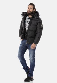 Schwarze Daunenjacke mit Reißverschluss, hohem Kragen und Logo-Detail; kombiniert mit blauen Jeans und schwarzen Turnschuhen. Modernes, funktionales Design.