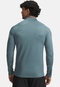 Camiseta atlética de manga larga en suave tela color azul verdoso con un patrón texturizado, cuello alzado y costuras en contraste. La vista trasera muestra un corte ajustado.