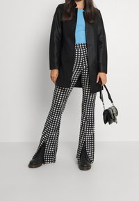 Abrigo de cuero sintético negro, blusa ajustada azul, pantalones acampanados en blanco y negro con estampado de pata de gallo, y botas negras con cordones, acompañados de un pequeño bolso negro.