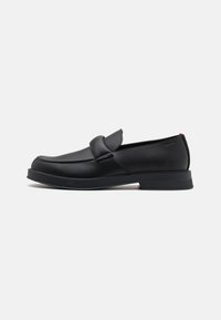 Mocassins en cuir noir avec une finition lisse, légèrement arrondie à l'avant, accent de sangle à l'avant et une semelle en caoutchouc texturée basse. Logo à l'arrière.
