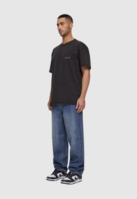 Svart bomulls t-skjorte med lilla logo, løs passform, kombinert med blå denimjeans og svart-hvite sneakers. Uformell gateklestil.