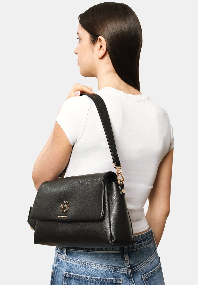 Mujer con cabello oscuro que lleva una camiseta blanca de manga corta y jeans, lleva un bolso de cuero negro con cierre dorado en el hombro.