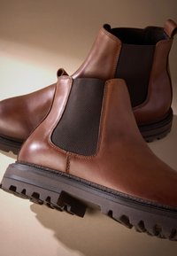 Hnedé kožené Chelsea topánky s elastickými bočnými panelmi, robustnými čiernymi gumovými podrážkami a detailným prešívaním. Hladká textúra, výška nad členok.