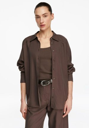 Frau trägt ein locker sitzendes braunes Hemd mit Knöpfen über einem passenden Tanktop, hoch taillierte Hose und schwarzen Gürtel, steht vor weißem Hintergrund.