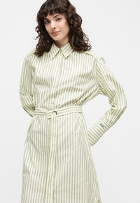 Νεαρή γυναίκα με σγουρά μαλλιά φοράει ένα κρεμ φόρεμα με λεπτές πράσινες κάθετες ρίγες, εμπρόσθιο κούμπωμα με κουμπιά, γιακά και ταιριαστή ζώνη.