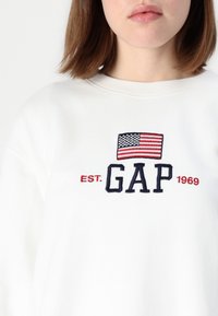 Sweatshirt blanc avec drapeau américain brodé et logo "GAP" en bleu marine. Le texte comprend "EST. 1969" en rouge sous le logo.