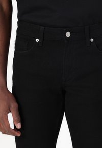 Jeans en denim noir avec quatre poches, une fermeture à bouton en métal et des détails de surpiqûres contrastants. Texture lisse et coupe droite.