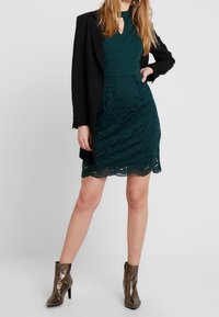 Grünes Spitzenkleid mit einem taillierten Oberteil und einem gewellten Saum, kombiniert mit einem schwarzen Blazer und metallischen Ankle Boots. Verfügt über komplizierte florale Muster.