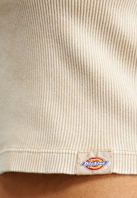 Beige ribbad tyg kännetecknar en åtsittande skjorta. Klämslutet visar en texturerad etikett med "Dickies"-logotypen. Mjuk textur, snug design.