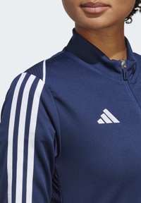 Navy Sportjacke mit hohem Kragen, Reißverschluss, strukturierten Seitenpaneelen und drei weißen Streifen auf jeder Schulter. Adidas-Logo auf der Brust.