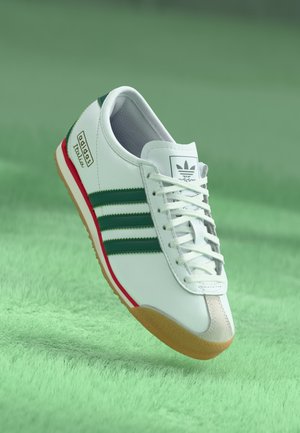 Chaussure Adidas Taekwondo blanche avec des bandes vertes, semelle en gomme et accents rouges flottant au-dessus d'une surface texturée verte.