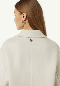 Witte wollen jas met een omgeslagen kraag, achternaad en een ronde zilveren knop met een logo. Het haar is steil en valt voorbij de schouders.