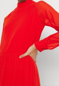 Pitkä punainen mekko, jossa on korkea kaulus ja pitkät, läpinäkyvät hihat. Hyvin laskostettu vyötärö ja sivutaskut. Sujuva pinta ja kirkas väri.