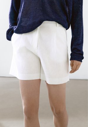 Person wearing white tailored shorts and a navy blue knit sweater, standing with one hand in a pocket against a neutral background.